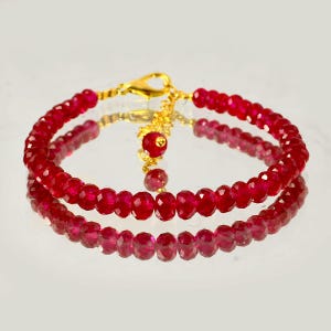 Pode incluir: Uma pulseira composta por pequenas contas facetadas de cor vermelho profundo enfiadas. A peça apresenta um fecho lagosta de metal dourado e uma corrente de extensão ajustável, terminada com uma única conta vermelha na extremidade.
