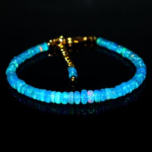 May include: A bracelet composed of small, round, vibrant blue opal beads with iridescent flashes of color. The bracelet features a gold-toned metal clasp and an adjustable chain extension, creating a circular shape against a dark background.