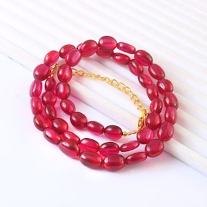 Puede incluir: Un collar que presenta cuentas de color rojo intenso, translúcidas y de forma ovalada. La pieza incluye un segmento de cadena de metal dorado con un cierre de mosquetón, lo que permite ajustar la longitud. Las cuentas tienen un acabado pulido.