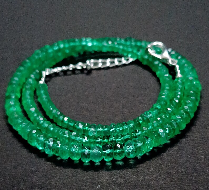 May include: A necklace composed of numerous small, faceted, vibrant green gemstone beads arranged in a circular pattern. The necklace features a polished silver-toned metal lobster claw clasp and a short length of silver-toned metal chain extender.
