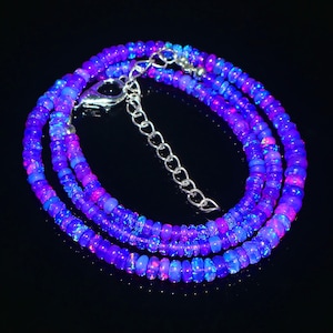 May include: A vibrant necklace composed of small, round, iridescent beads in shades of blue, purple, and pink. The beads are strung together, forming multiple strands. A silver-colored clasp and extender chain complete the necklace, which is set against a black background.