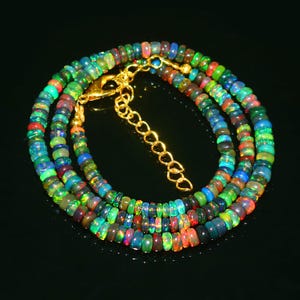 May include: A multi-colored opal bead necklace with a gold-colored clasp and extender chain. The beads display a range of colors, including green, blue, red, and black. The necklace is displayed on a black surface.