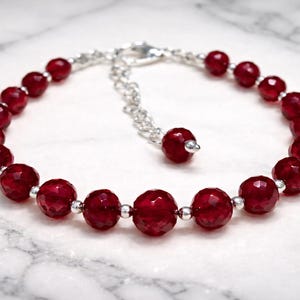 Pode incluir: Uma pulseira com contas facetadas em vermelho profundo alternadas com pequenas esferas de metal prateado. A peça inclui uma corrente de extensão de metal prateado com uma única conta vermelha na extremidade e um fecho tipo lagosta.