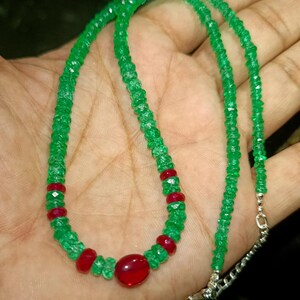 May include: A necklace featuring a strand of small, faceted green beads interspersed with several smooth, round red beads. The necklace includes a silver-toned metal chain extension with a clasp at the end, creating a vibrant contrast of colors.