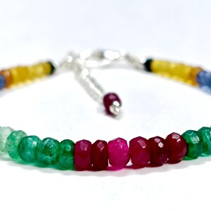 Könnte beinhalten: Ein mehrfarbiges Perlenarmband mit kleinen, facettierten, runden Edelsteinen in den Farben Rot, Grün, Blau und Gelb. Das Armband verfügt über einen silberfarbenen Metallverschluss und eine Verlängerungskette mit einer einzelnen roten Perle am Ende.
