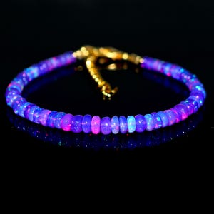 May include: A beaded bracelet with iridescent blue, purple, and pink opal stones. The bracelet has a gold-colored clasp and is set against a black background. The opal beads are round and evenly sized.
