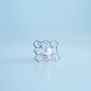 Op de afbeelding: Een zilverkleurige ring met een geometrisch ontwerp, met een centrale witte parel. Het openwerkpatroon van de ring creëert een honingraatachtige structuur. De achtergrond is zacht, lichtblauw.
