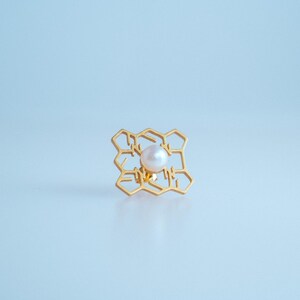 May include: A gold-colored ring with a honeycomb design, centered with a white pearl. The ring is set against a light blue background, creating a minimalist aesthetic. The ring's intricate design is a striking feature.