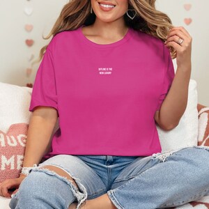 Op de afbeelding: Een vrouw draagt een roze T-shirt met de tekst "OFFLINE IS THE NEW LUXURY" in wit. Ze zit op een witte bank met een roze hartvormig kussen waarop "HUG ME" staat.