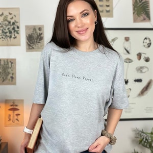 Può includere: T-shirt grigio chiaro con la scritta "Code. Sleep. Repeat." in nero. La maglietta ha una vestibilità ampia e maniche corte. Un capo casual per l'uso quotidiano.