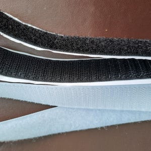 May include: A strip of black hook-and-loop fastener tape featuring a textured, fuzzy surface on one side and a rigid, hooked surface on the other, with a white adhesive backing visible along the edges of the black material.