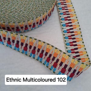 Op de afbeelding: Close-up van een rol kleurrijke geweven bies. De bies heeft een herhalend patroon van rode, oranje, gele en blauwe rechthoeken op een cr&egrave;mekleurige achtergrond. De tekst "Ethnic Multicoloured 102" is zichtbaar.