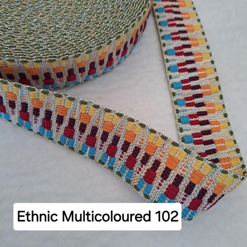 Op de afbeelding: Close-up van een rol kleurrijke geweven bies. De bies heeft een herhalend patroon van rode, oranje, gele en blauwe rechthoeken op een cr&egrave;mekleurige achtergrond. De tekst "Ethnic Multicoloured 102" is zichtbaar.