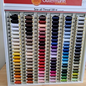 May include: A wall display of Gutermann Sew-all Thread in 100 metre spools. The spools are arranged in rows by colour, with a variety of colours including white, black, brown, red, pink, purple, blue, green, and grey.