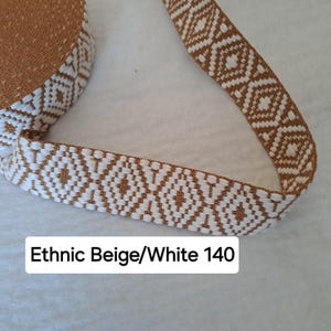 Op de afbeelding: Close-up van een beige en wit patroonlint. Het lint heeft een herhalend geometrisch diamantontwerp. De tekst "Ethnic Beige/White 140" is zichtbaar. Dit lint is geschikt voor knutsel- of naaiprojecten.