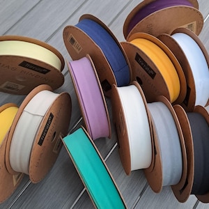 May include: A selection of bias binding spools in a range of colours, such as yellow, blue, purple, white, and teal. The spools are made of cardboard and are arranged on a grey wooden surface. The text "BIAS BINDING" is printed on the spools.