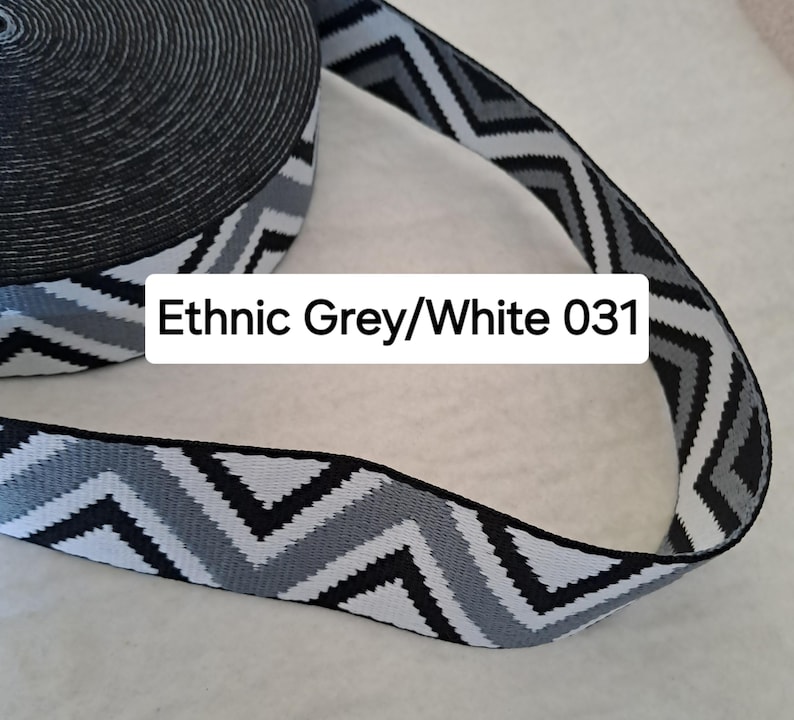 Op de afbeelding: Een rol elastisch lint met een zwart-wit patroon en een zigzagontwerp. Het lint heeft een herhalend patroon van zwarte, grijze en witte zigzags. De tekst "Ethnic Grey/White 031" staat op het lint.