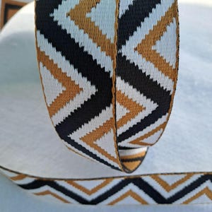 Op de afbeelding: Close-up van een decoratief lint met een herhalend chevronpatroon. Het lint heeft een ontwerp van zwarte, witte en gouden chevrons. Het lint is opgerold, met een deel afgerold aan de onderkant, wat de continu&iuml;teit van het patroon laat zien.