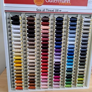 May include: A wall display of Gutermann Sew-all Thread in 100 metre spools. The spools are arranged in rows by colour, with a variety of colours including white, black, brown, red, pink, purple, blue, green, and grey.