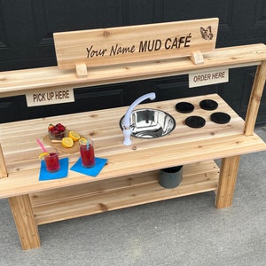 May include: A wooden play kitchen with a stainless steel sink, a tap, and a sign that reads "Your Name MUD CAFÉ". The kitchen has a lower shelf and a sign that says "PICK UP HERE". Drinks and fruit are on the counter.
