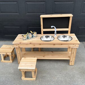 May include: A wooden outdoor play kitchen set with two stainless steel bowls, a tap, and a chalkboard. The set includes two small wooden stools and a tray with utensils. The wood has a natural finish.