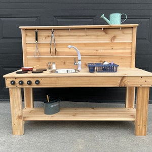 May include: A wooden outdoor play kitchen with a sink, tap, and hob. Includes play accessories like a pan, utensils, and a watering can. The kitchen is made of light-coloured wood and has a lower shelf for storage.