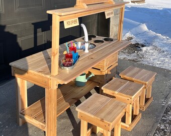 May include: A wooden mud kitchen set with a sink, stove, and serving area. The set includes three matching stools. The sign reads "Your Name MUD CAFE". The set is made of light-colored wood and is designed for outdoor play.
