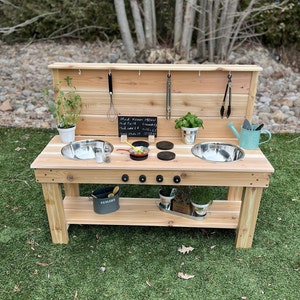 May include: A wooden outdoor play kitchen in light brown. It has two stainless steel sinks, a small chalkboard menu, and various cooking utensils. A small watering can and potted plants are also included, adding to the kitchen's appeal.