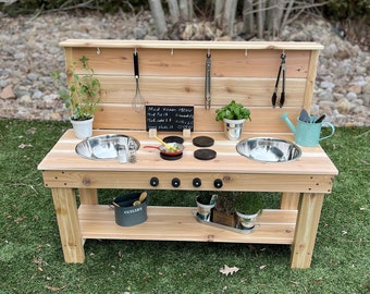 May include: A wooden outdoor play kitchen with a light brown finish. It features two stainless steel sinks, a small chalkboard menu, and various cooking utensils. A small watering can and potted plants add to the play kitchen's charm.