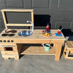 May include: A wooden play kitchen set with a sink, stove, oven, and chalkboard. The set includes a small stool and a watering can. The kitchen is made of light-coloured wood and has a stainless steel sink.