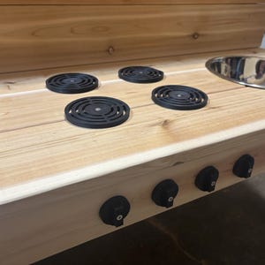 Könnte beinhalten: Eine Spielküche aus Holz mit hellbraunem Finish. Sie verfügt über zwei Edelstahl-Schalen, vier schwarze Kochfeldabdeckungen und schwarze Knöpfe. Die Spielküche ist für Kinder konzipiert.