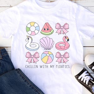 Könnte beinhalten: Weißes T-Shirt mit bunten Cartoon-Illustrationen von Schwimmtieren, darunter ein Flamingo und ein Einhorn. Der Text lautet "CHILLIN WITH MY FLOATIES". Enthält auch blaue Jeans und schwarze Turnschuhe.