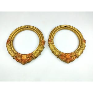 May include: Two ornate, gold-colored, circular frames with decorative detailing. The frames have a weathered appearance with reddish-orange accents on the lower portion. Each frame has a small hole at the top for hanging. The frames are set against a white background.