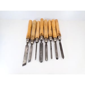May include: A set of eight wood carving chisels and gouges with light-colored wooden handles and metal ferrules. The tools feature various blade shapes, including flat, angled, and curved edges, all showing signs of use with metallic surfaces.