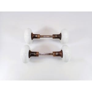 May include: Two sets of door knobs featuring smooth, round white porcelain handles attached to dark, weathered metal shanks. Each assembly includes a central metal spindle connecting the two knobs, secured by small flathead screws on the metal collars.