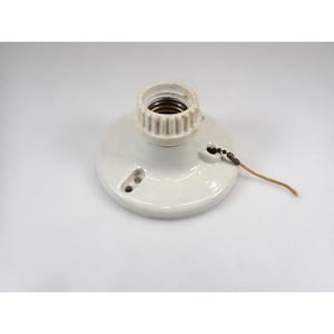 May include: A vintage white ceramic light socket with a brass interior. The socket has a round base with two small holes and a pull chain. The electrical component is designed for a standard light bulb.