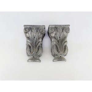 May include: A pair of matching grey wall corbels featuring a weathered, textured finish and an ornate acanthus leaf design. Each architectural bracket has a flared top, a tapered body, and a small circular mounting hole at the top and bottom.
