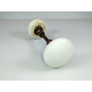 May include: Two white oval-shaped porcelain door knobs connected by a metal rod. The knobs have a slightly aged appearance with subtle discoloration. The metal components show signs of age and wear, suggesting a vintage aesthetic.