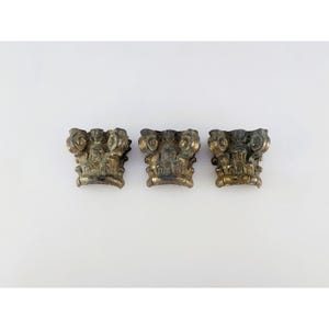 May include: Three small, metallic, gold-toned decorative objects shaped like architectural column capitals. Each piece features a curved, scroll-like design at the top and a flared base with intricate, weathered detailing and dark patina accents.