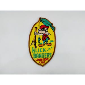 May include: An oval-shaped embroidered patch with a yellow background and a brown border. It features a cartoon character wearing a red cap, red vest, and blue pants, holding a fishing rod and a rifle. The text reads KLICK-M-BANGERS LEMON GROVE CALIF.