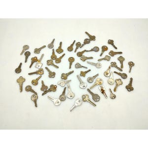 Könnte beinhalten: Eine Sammlung von Vintage-Schlüsseln in verschiedenen Formen und Größen, verstreut auf einer weißen Oberfläche. Die Schlüssel sind hauptsächlich metallisch, mit Schattierungen von Silber, Bronze und Messing. Einige Schlüssel weisen Alters- und Abnutzungserscheinungen auf.