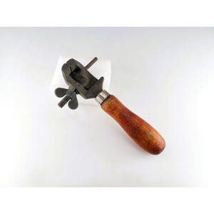 May include: A vintage metal hand vise tool featuring a dark, weathered iron clamp mechanism with a threaded wing nut adjustment screw and a smooth, cylindrical wooden handle with a warm brown finish, showing signs of age and consistent use.