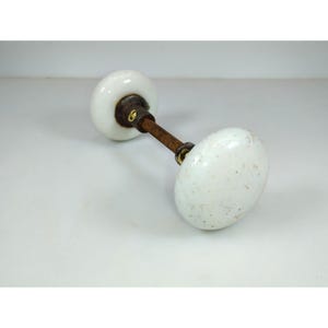 May include: A pair of antique white porcelain door knobs with a speckled pattern. The knobs are connected by a rusty metal rod and brass hardware. The knobs are oval-shaped and have a classic, vintage aesthetic.