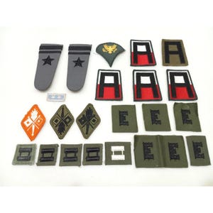 May include: A collection of military patches and insignia, including shoulder boards with stars, patches with the letter "A" and "E", and various rank insignia. The patches are in shades of gray, red, green, and white.