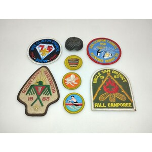 May include: A collection of vintage Boy Scout patches and pins. The patches feature various designs, including a diamond with text, a canoe, a campfire, and a Native American-inspired emblem. The patches are in various colors, including blue, green, and yellow.
