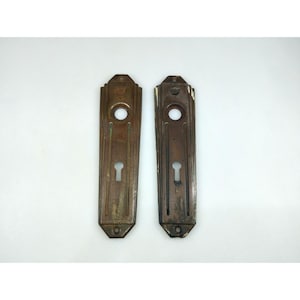 May include: Two antique bronze door plates with keyholes and circular handle openings. The rectangular plates have vertical grooves and decorative tops. The metal has a weathered appearance with areas of discoloration.