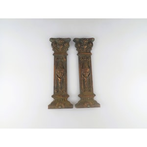 May include: A pair of vertical rectangular bronze-toned metal architectural elements featuring ornate capitals at the top and a central relief carving of a human face wearing a pointed headpiece, with decorative scrollwork at the base.