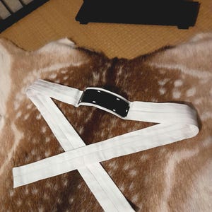 Puede incluir: Un cinturón obi blanco con una pieza central negra y un objeto de madera en blanco y negro en el fondo. El cinturón está hecho de una tela blanca suave y está diseñado para atarse a la cintura. La pieza central negra tiene costuras blancas.