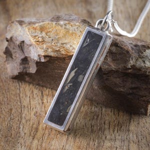 May include: A rectangular pendant necklace with a silver chain. The pendant features a dark, textured stone set in a silver frame. The necklace is displayed on a piece of rough rock against a wooden background. The text "VESTIS STUDIOS" and "TIMELESS COLLECTION" are visible.