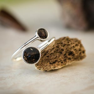 Puede incluir: Anillo de plata con dos piedras circulares oscuras. La banda del anillo se curva alrededor del dedo, con una piedra colocada encima de la otra. Parte de la colección Timeless de Vestis Studios.